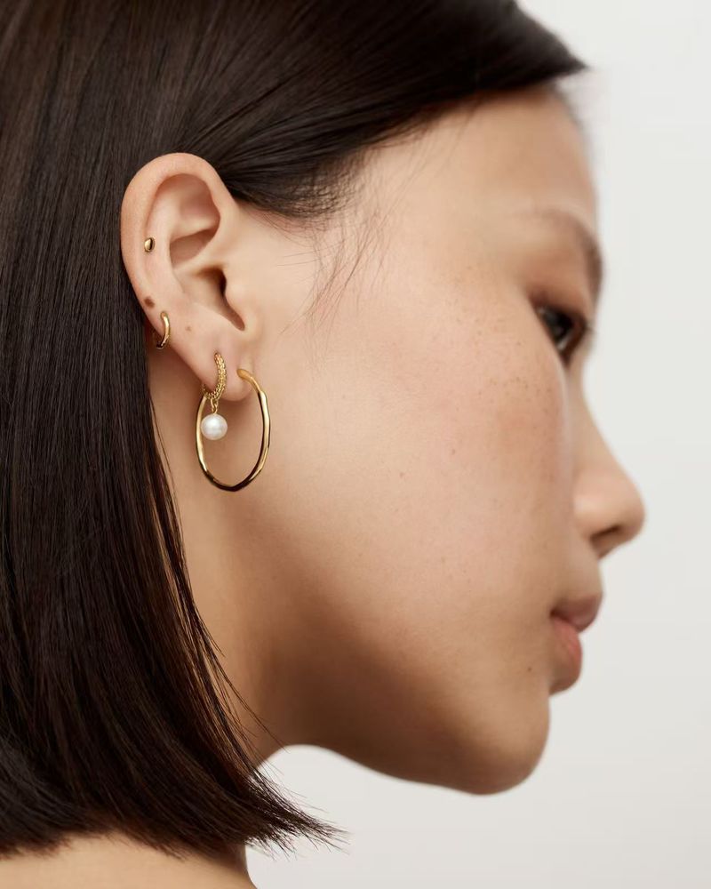 Close-up profile of a woman wearing gold hoop earrings with a small white pearl charm against a plain light background