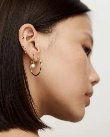 Close-up profile of a woman wearing gold hoop earrings with a small white pearl charm against a plain light background