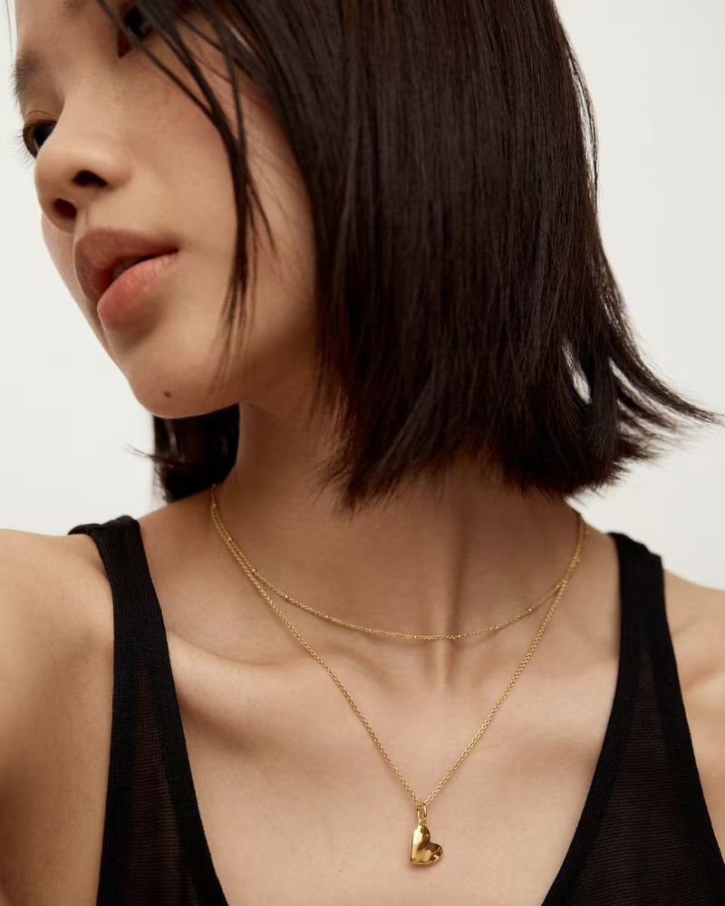 Close-up shot of a young Asian woman wearing a gold necklace with a heart pendant, against a neutral background in soft lighting.