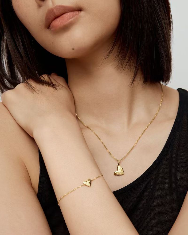 Close-up of a woman wearing a gold heart pendant necklace and matching heart bracelet against a pale neutral background.