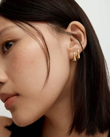 Close-up studio shot of an Asian woman with dark hair, wearing a pair of modern gold earrings, against a neutral background.