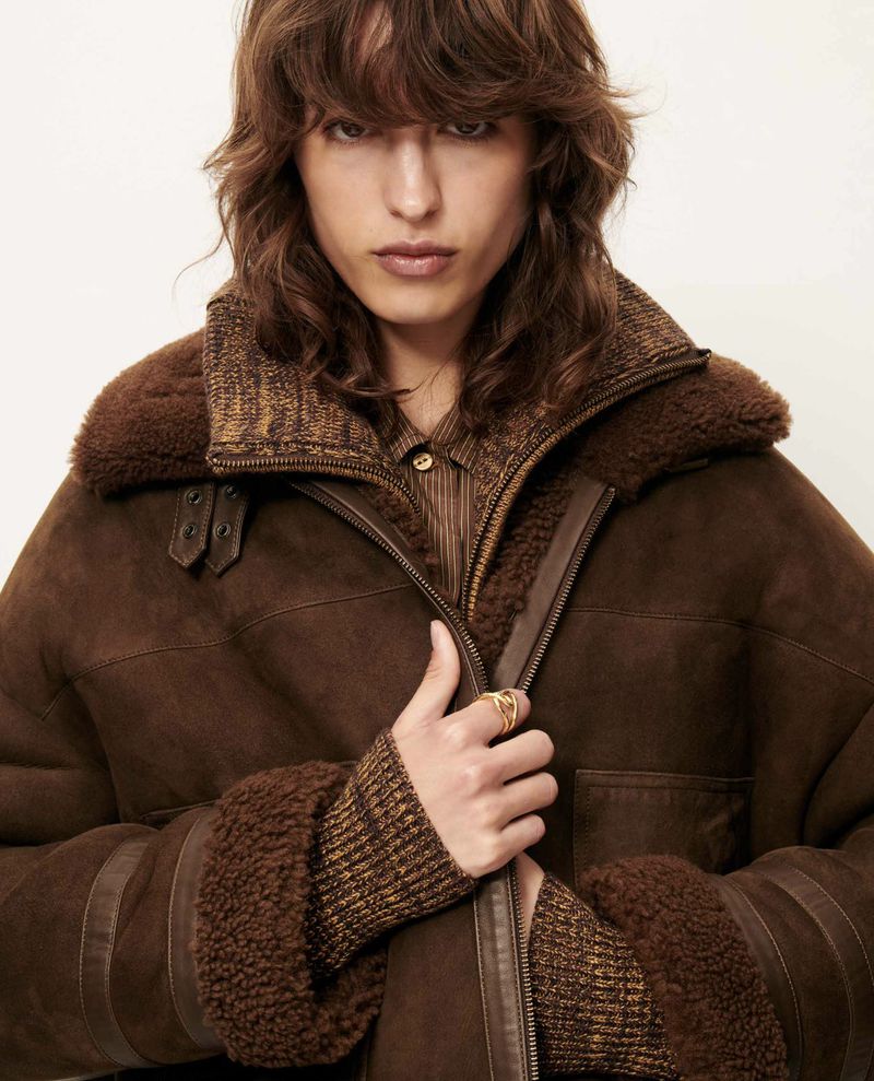 Close-up of adult wearing a brown shearling bomber jacket and brown striped top against a white background in a studio setting.