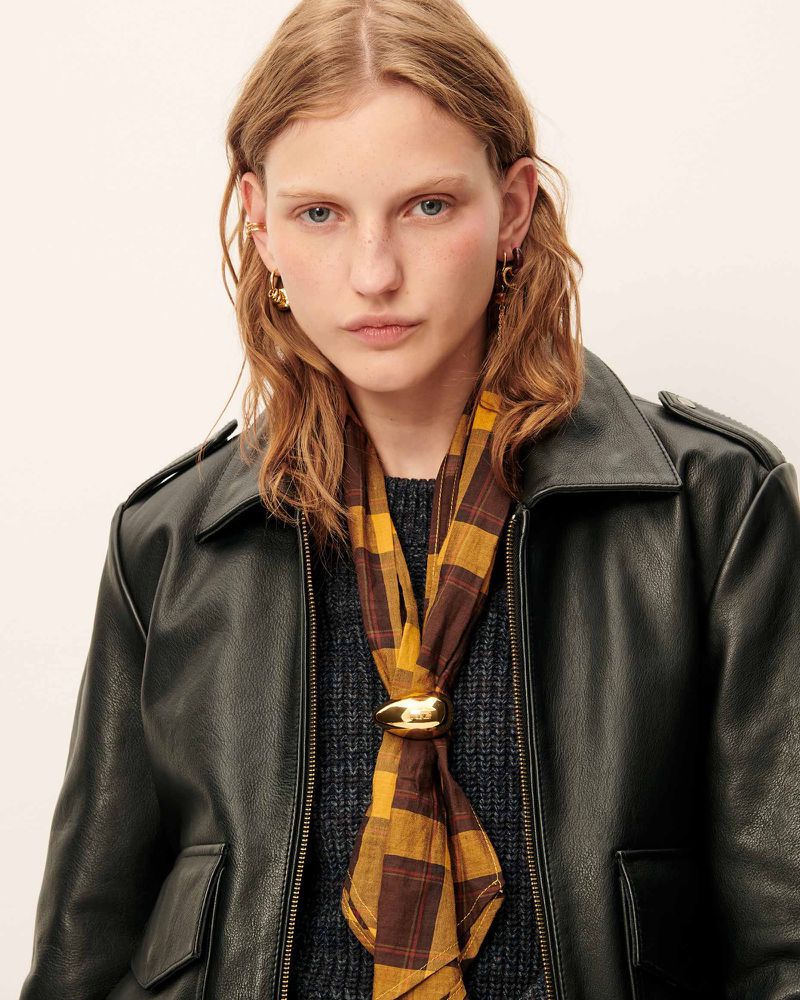 Blonde woman wearing black leather jacket and plaid scarf, posing indoors, eye-level. Neutral expression. Fashion photography.