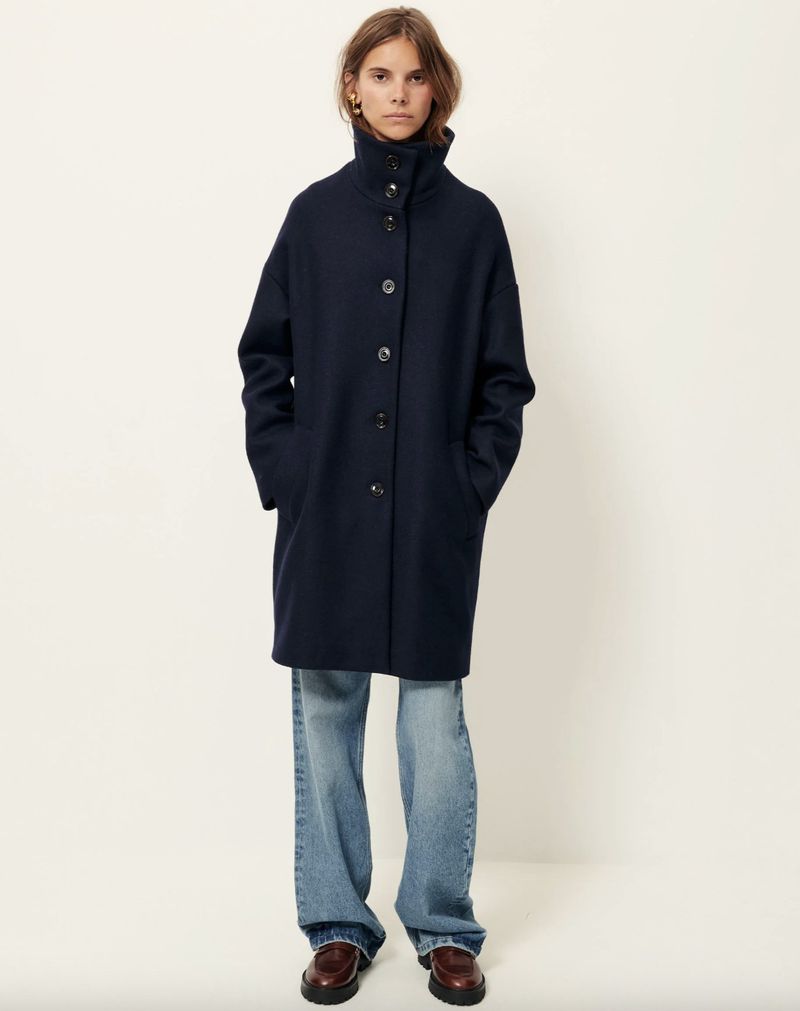 A woman stands in a studio wearing a navy wool coat, light blue jeans, and brown loafers against a bright, neutral background.