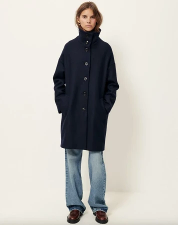 A woman stands in a studio wearing a navy wool coat, light blue jeans, and brown loafers against a bright, neutral background.