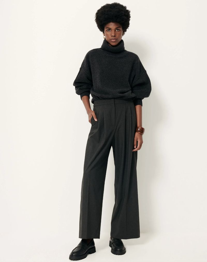 Dark-skinned woman with dark, natural afro poses in a studio wearing a dark turtleneck sweater, black slacks, and black shoes.