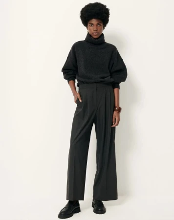 Dark-skinned woman with dark, natural afro poses in a studio wearing a dark turtleneck sweater, black slacks, and black shoes.