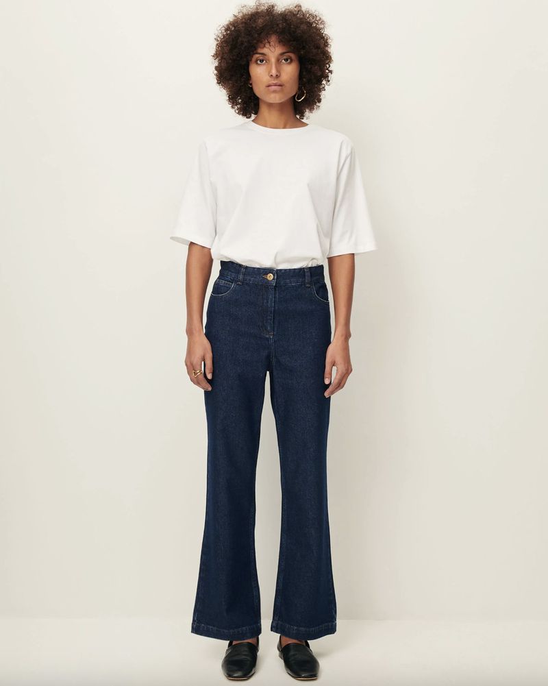 A woman in a white T-shirt and dark wash, wide-leg denim jeans poses in front of a white backdrop in a fashion forward shot.