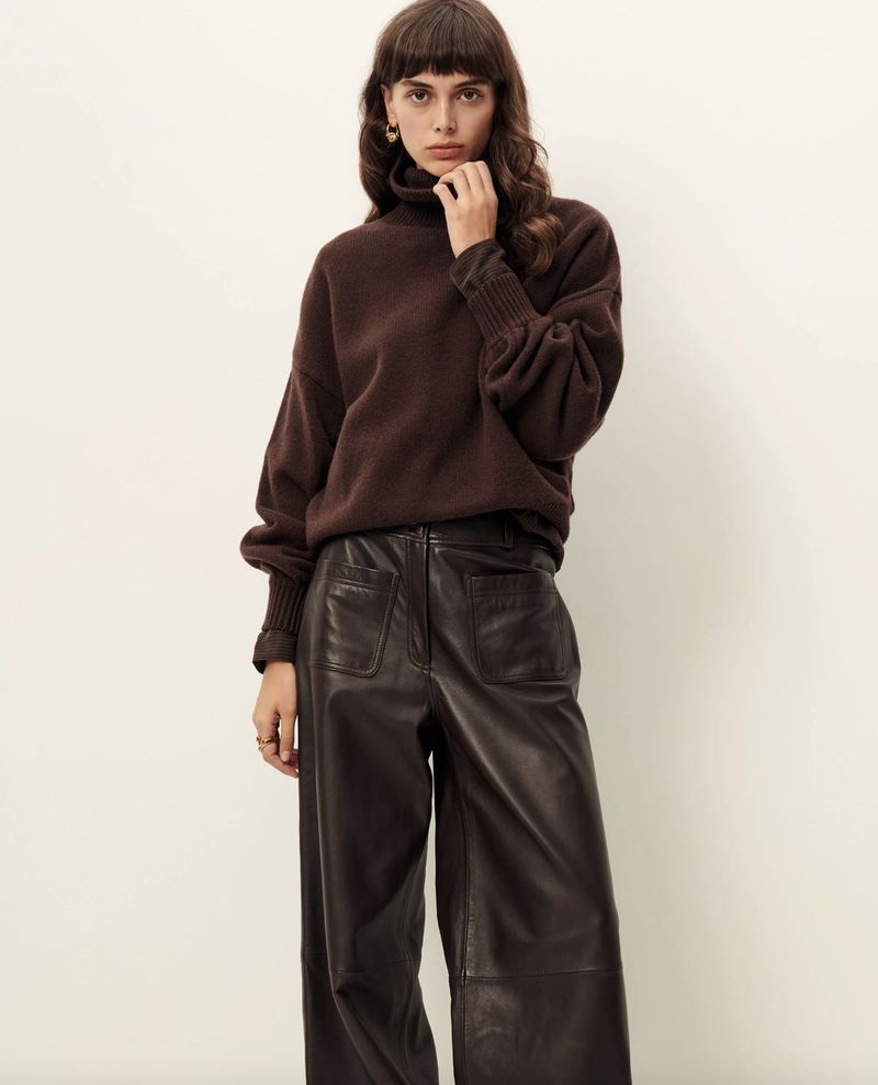 Woman wearing a brown turtleneck sweater and wide-leg leather pants in a studio setting, fashion photography, full shot, neutral backdrop.