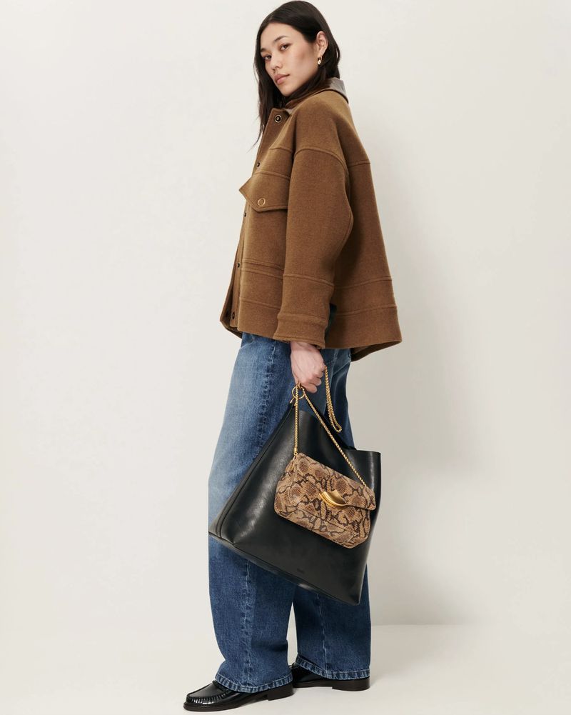 Studio shot of a female model wearing a brown jacket and blue jeans, holding a black tote bag with a patterned purse.
