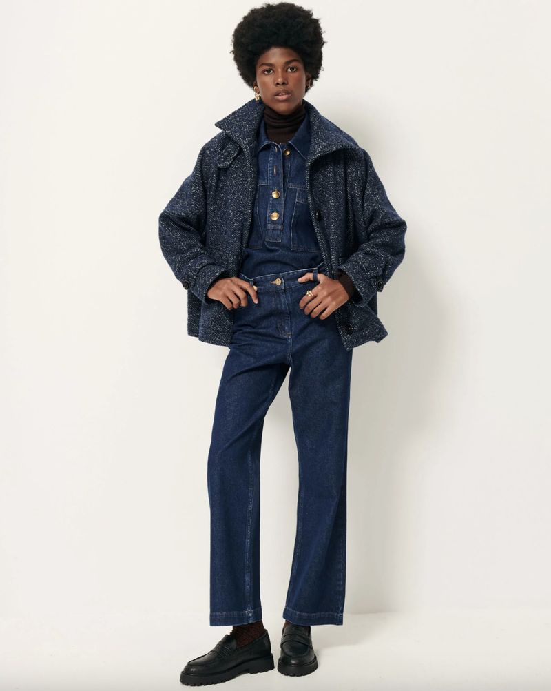 Full-length shot of a woman with an afro hairstyle wearing a denim jumpsuit, a jacket, and platform loafers against a white background.