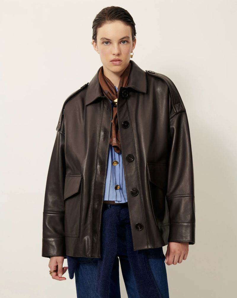 Adult female wearing a dark brown leather jacket, blue jeans and a printed scarf, posing in a bright indoor photo studio.