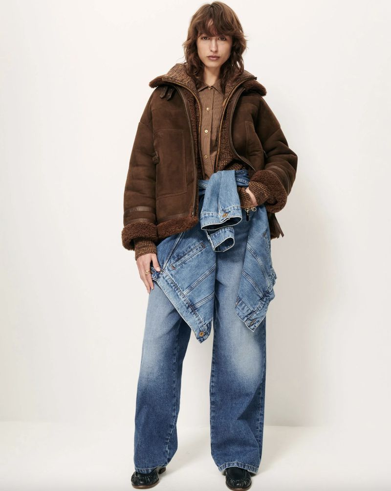 Full-length portrait of a woman wearing a brown suede jacket, brown shirt, and baggy jeans, against a white backdrop in a fashion editorial.