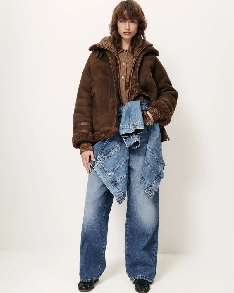 A woman poses in an indoor setting, wearing a brown jacket, denim bottoms and black shoes against an off-white backdrop.