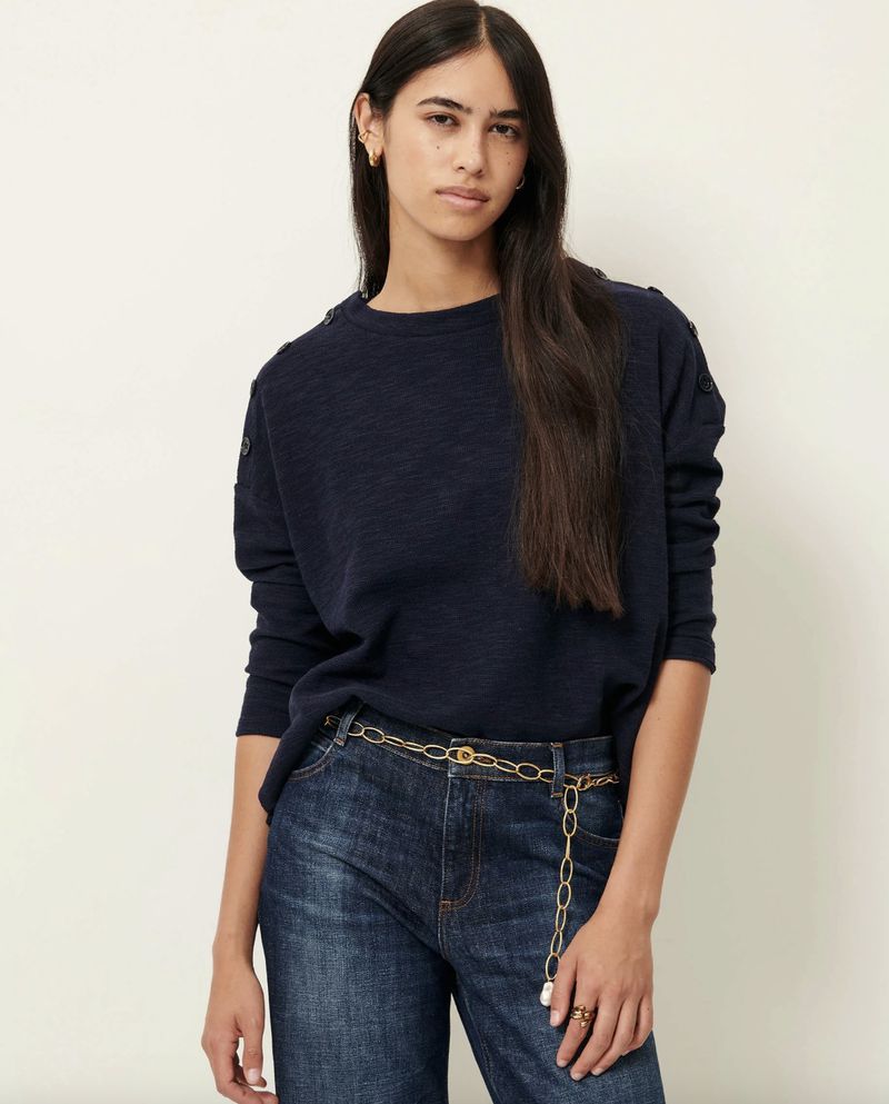 A brunette woman in a navy blue long-sleeve shirt and jeans poses in front of a neutral studio background, showcasing casual fashion.