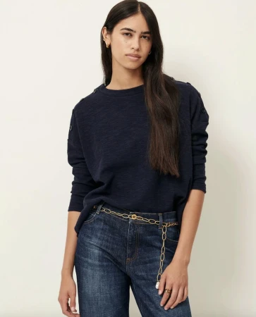 A brunette woman in a navy blue long-sleeve shirt and jeans poses in front of a neutral studio background, showcasing casual fashion.