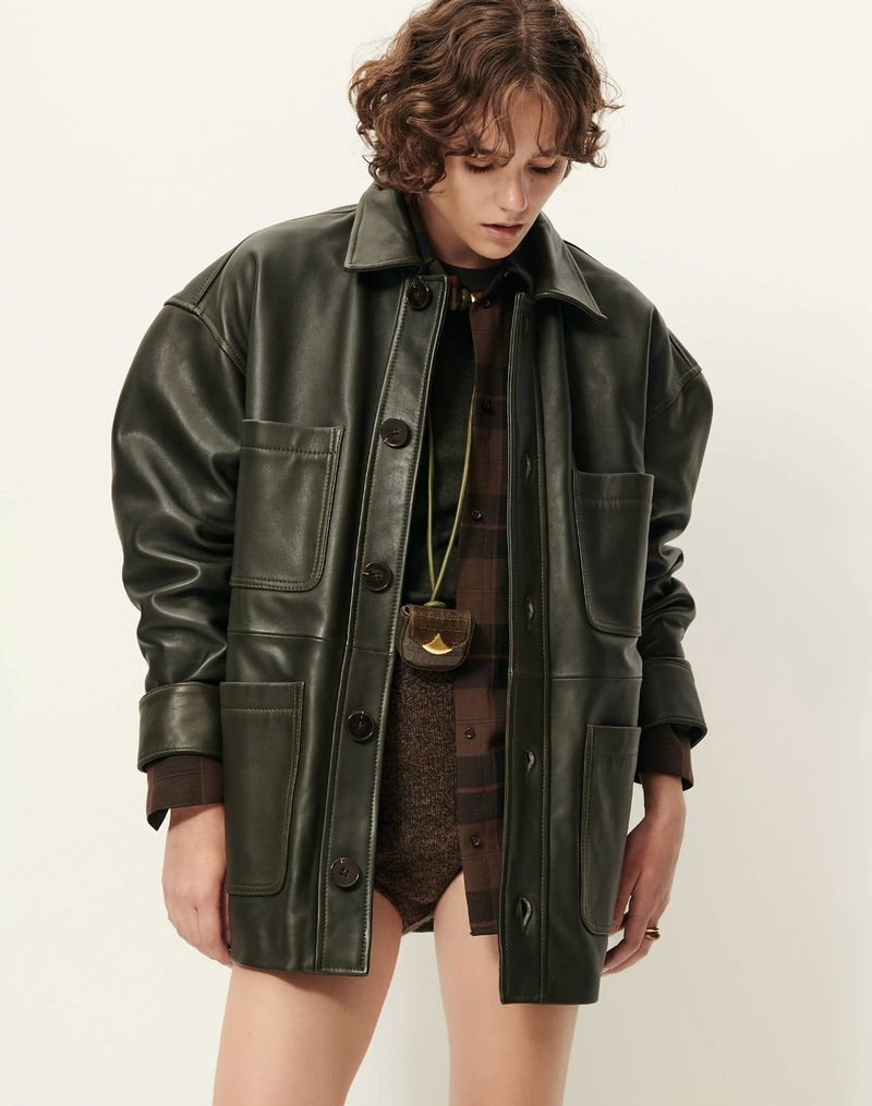 Fashion shot of a model wearing an olive green leather jacket and brown patterned top, posing against an off-white backdrop in a studio.