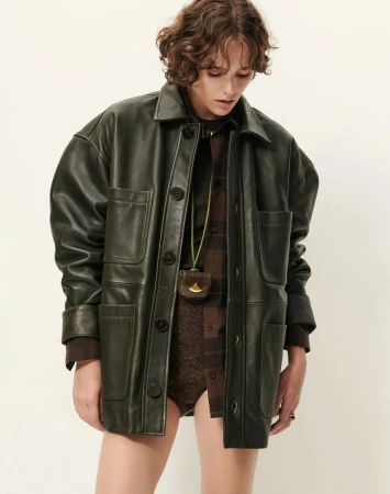 Fashion shot of a model wearing an olive green leather jacket and brown patterned top, posing against an off-white backdrop in a studio.