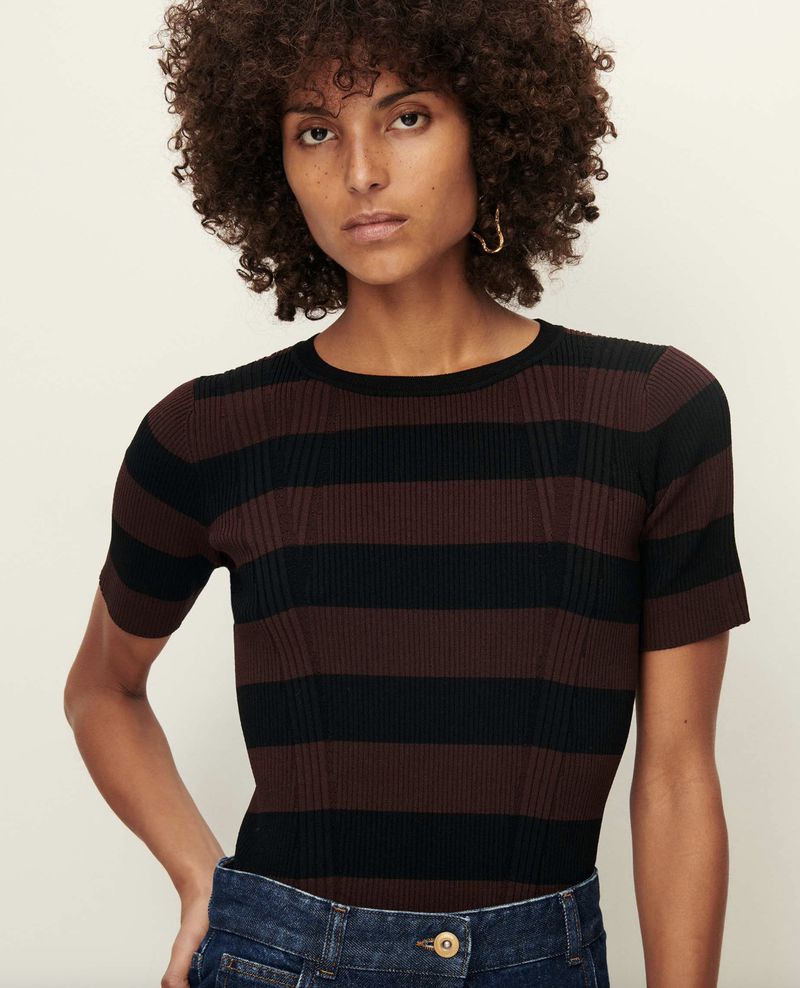 Adult female poses in a studio wearing a brown and black striped, short-sleeved shirt and jeans against a light background.