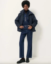 Model with an afro hairstyle posing in a dark blue denim set, speckled navy jacket, and black loafers on a white backdrop.