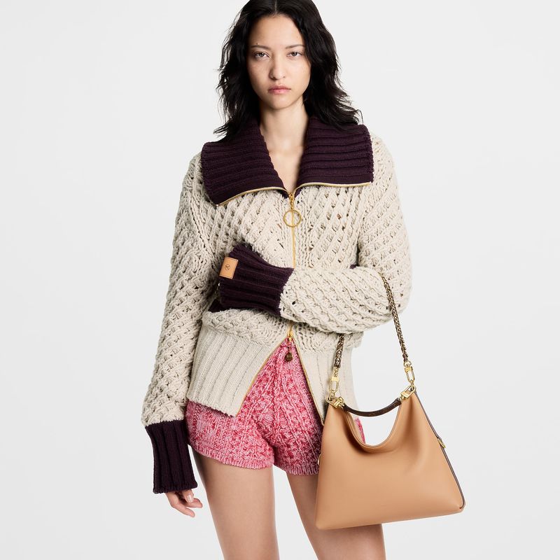 Asian fashion model poses in a cream knit zip-up sweater with a tan handbag and pink knit shorts on a white background.