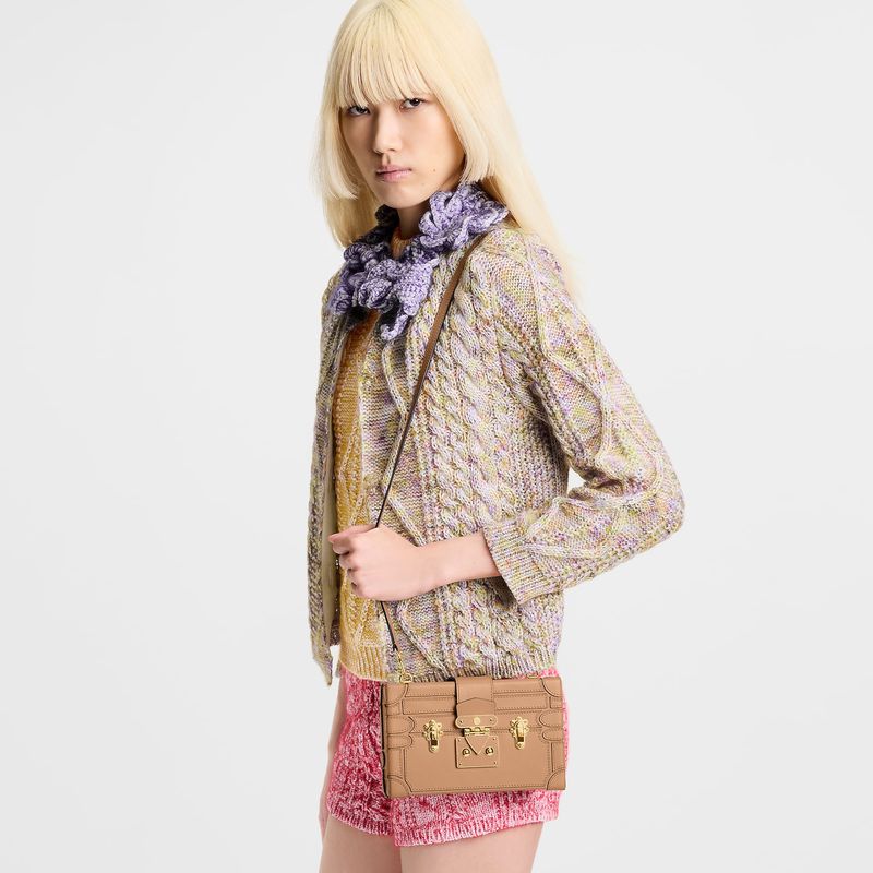 A young, fair-skinned woman wearing a knitted sweater, lilac scarf, and pink knit shorts with a brown leather purse on a white background.