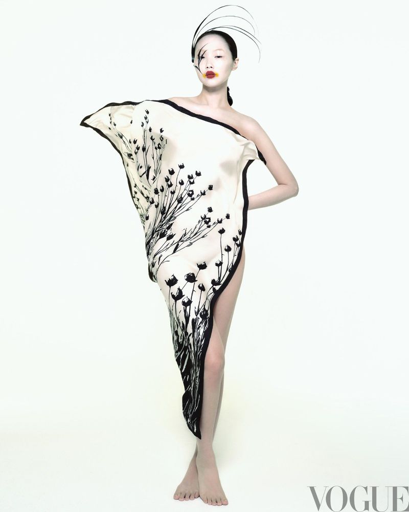 Asian woman in avant-garde fashion wearing a floral one-shoulder dress and feathered headpiece against a white background. Vogue logo.