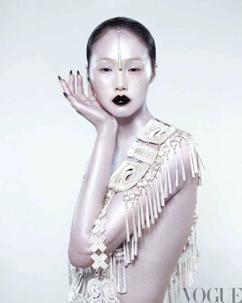 Striking fashion portrait of an Asian woman with glossy pale skin and black lips, adorned with ornamental shoulder piece against a white backdrop.