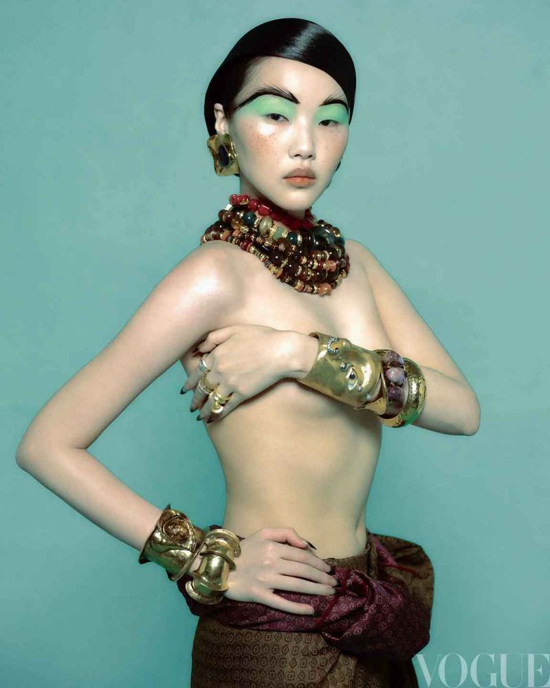 Elegant woman with bold makeup and jewelry poses in front of a turquoise background, featuring gold cuffs and a traditional skirt.