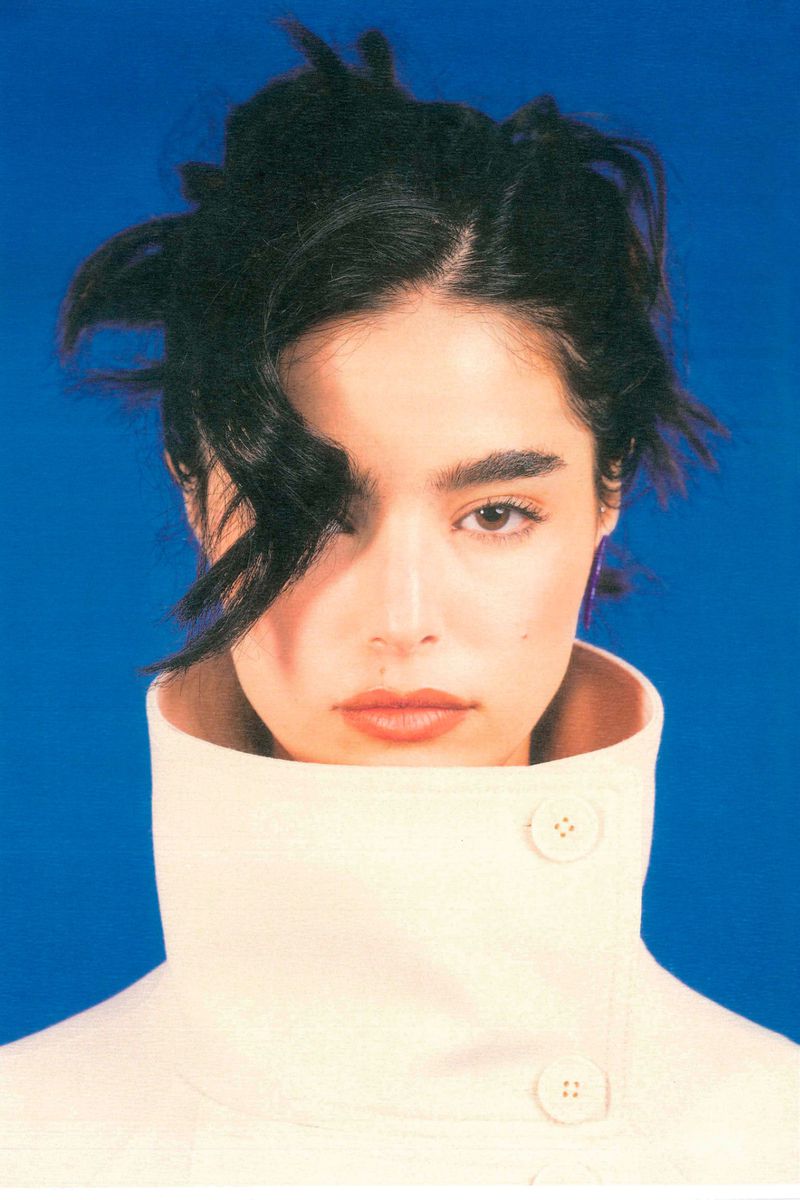Close-up portrait of a young woman with dark hair and light skin wearing a collared cream-colored jacket against a blue background.