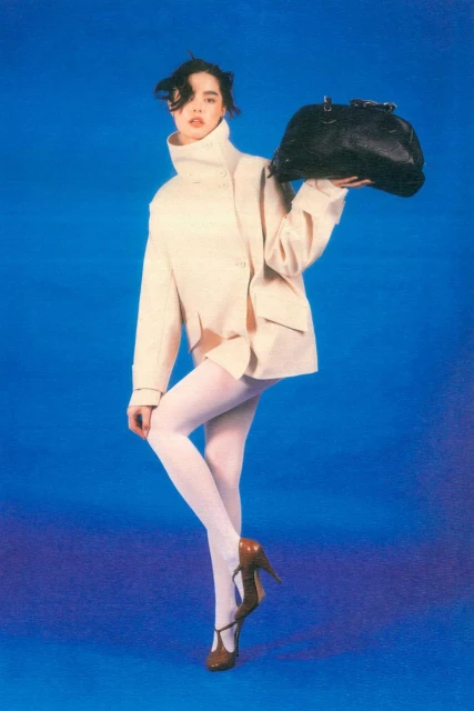 A slender fashion model poses in a studio against a blue background, wearing a cream-colored jacket, white tights, brown heels, and holding a black bag.
