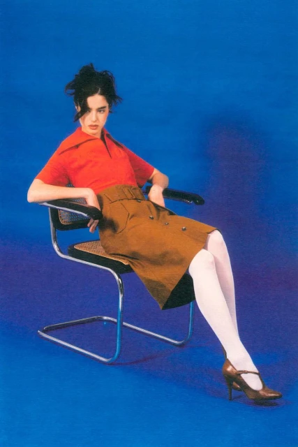 A young woman in a vintage orange top and brown skirt poses seated in a modern chair against a bright blue backdrop in a studio setting.
