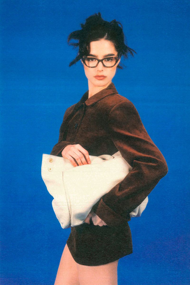 Portrait of a woman in a brown jacket and glasses, holding a white bag, against a solid blue background in a studio setting.