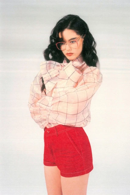 Young woman in red shorts and a sheer, patterned blouse poses indoors against a bright background. The portrait emphasizes fashion and style.