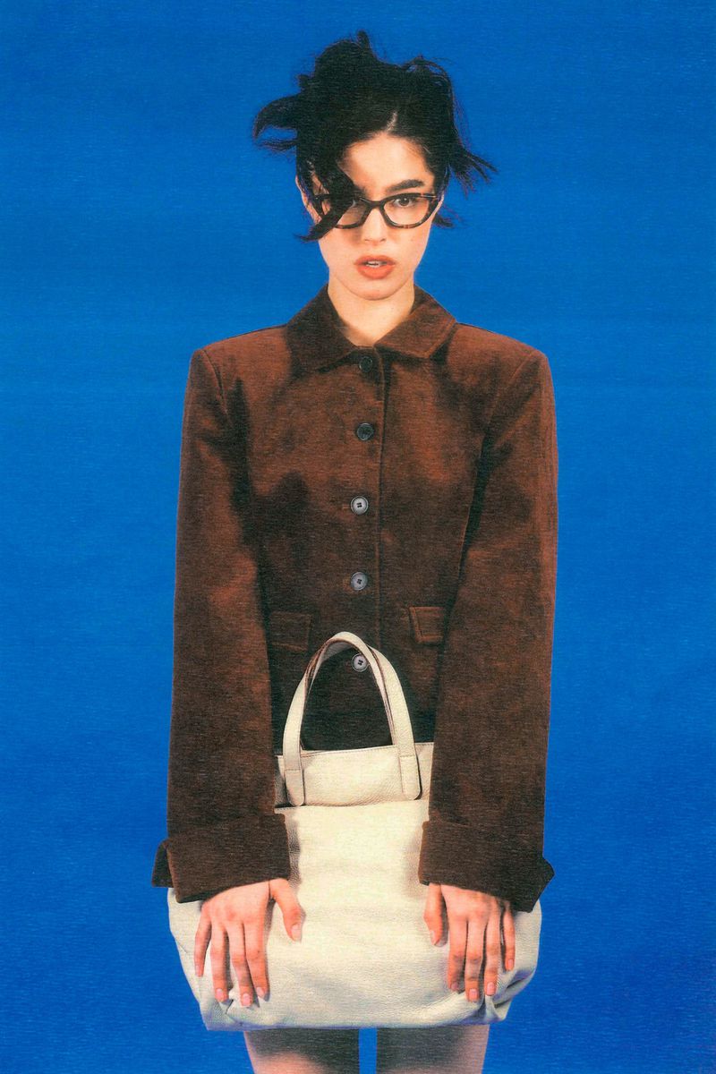 A young woman with messy dark hair and glasses poses in a brown jacket and skirt, holding a large white tote bag against a bright blue backdrop.