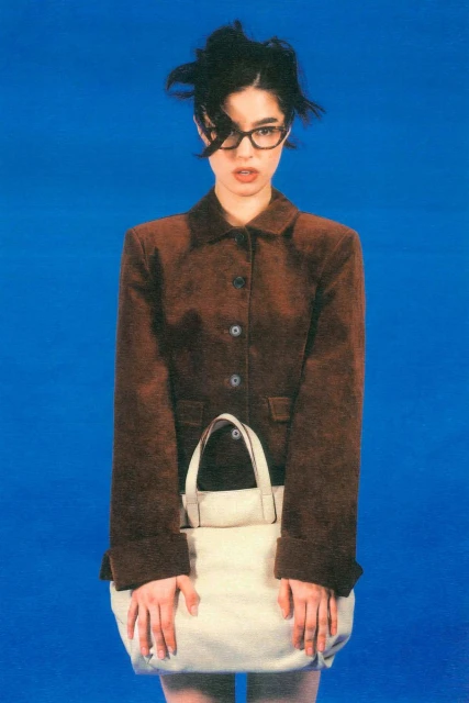 A young woman with messy dark hair and glasses poses in a brown jacket and skirt, holding a large white tote bag against a bright blue backdrop.