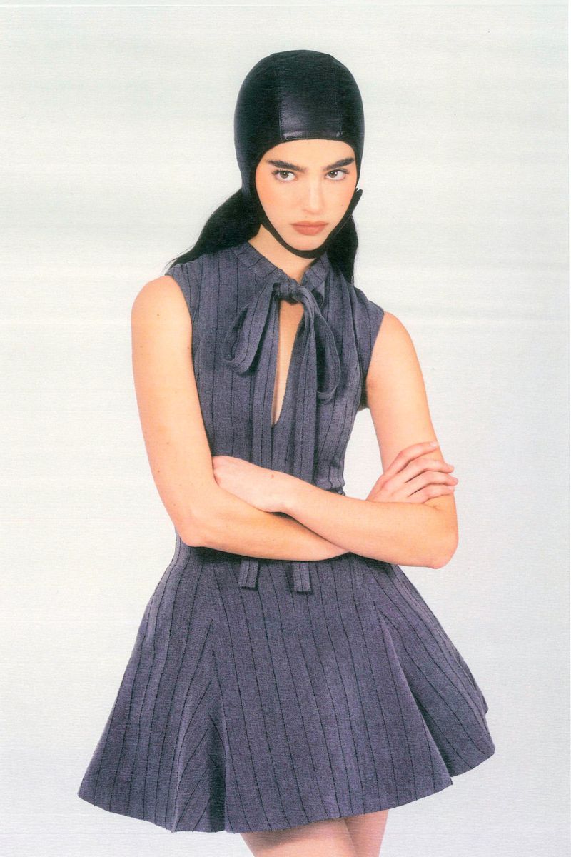 Fashion shot of an adult female wearing a gray striped dress with a black leather cap, arms crossed, and a neutral expression against white.