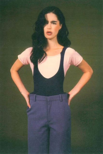 A young adult woman in a retro outfit poses with hands on hips against a green background, looking directly at the camera.
