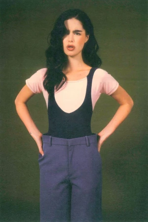 A young adult woman in a retro outfit poses with hands on hips against a green background, looking directly at the camera.