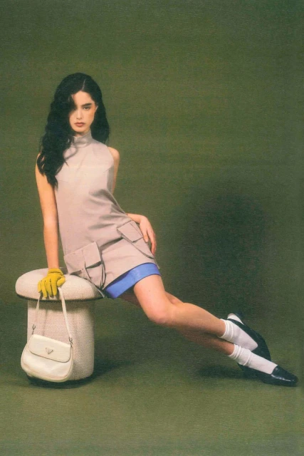 A brunette woman poses seated on a mushroom-shaped stool with a handbag, wearing a sleeveless dress, white socks, and black Mary Jane shoes.
