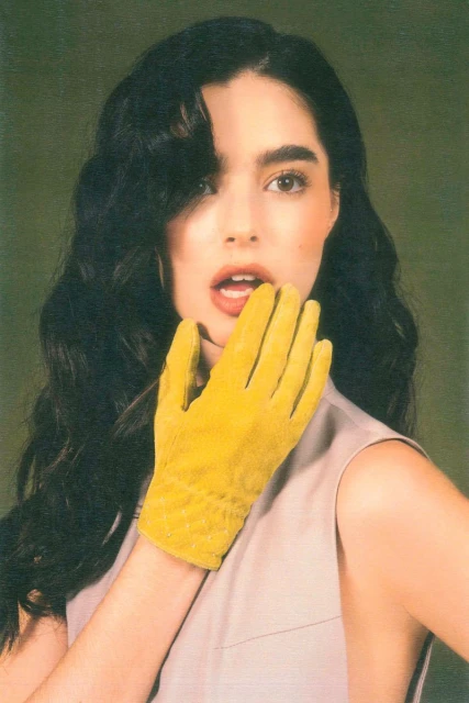 A woman with dark hair wears a pale tank top and yellow glove, looking directly at the camera in a portrait shot against a green background.