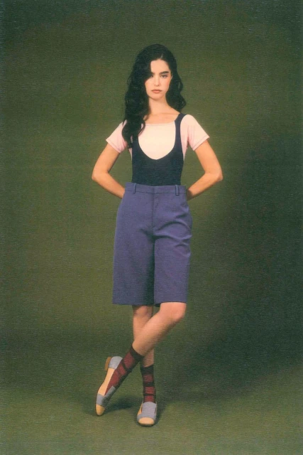 A young woman with long dark hair poses in a pink t-shirt, black suspender top, blue shorts, striped socks and flats against an olive backdrop.