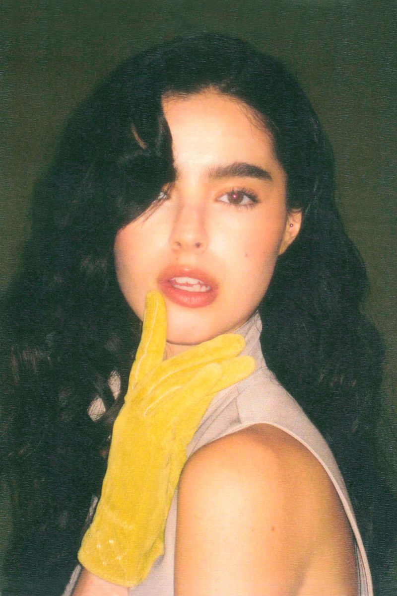 Woman with dark hair poses indoors, wearing a high-necked top and a yellow glove touching her lips, creating a striking beauty shot.