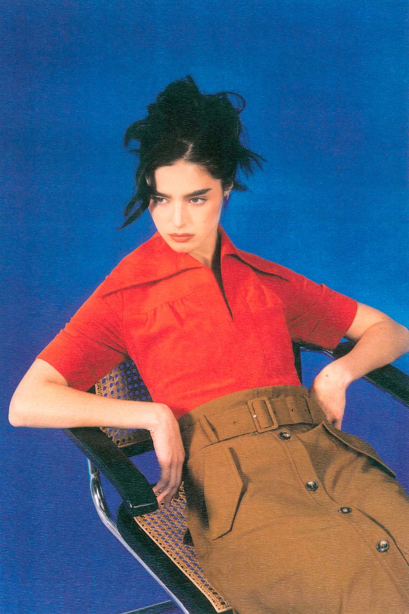 Elegant woman in a red collared shirt and brown skirt poses in a black chair against a vibrant blue background, exuding a retro fashion vibe.