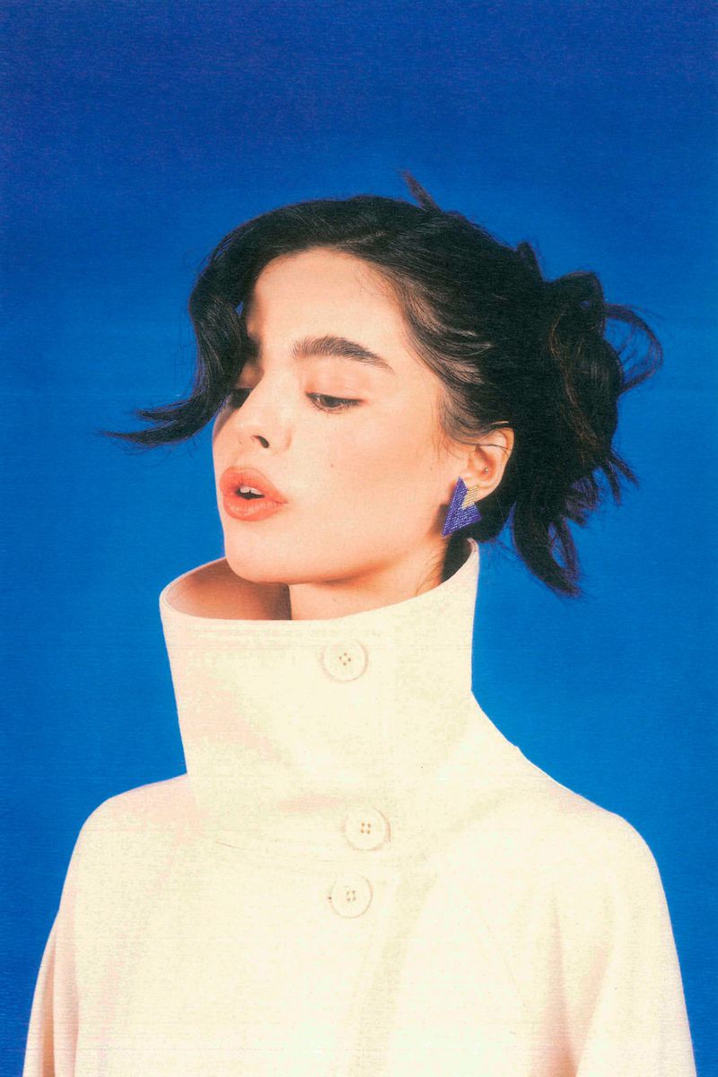 Portrait of a young adult woman with dark hair in a cream-colored coat with a high collar, against a bright blue background.
