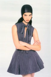 Model wearing a grey sleeveless bow dress and black headpiece with crossed arms against a pale studio backdrop.