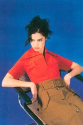 A woman in an orange collared top and belted camel skirt sits in a cane back chair against a bright blue studio backdrop.