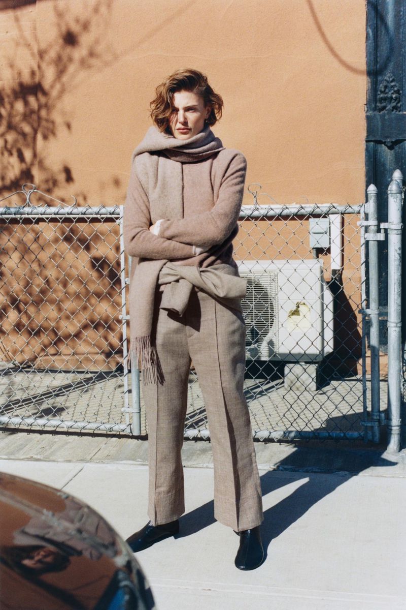 Model Bibi Breslin standing on a sunny sidewalk in a beige wool coat, matching trousers, and a wrapped fringed scarf.