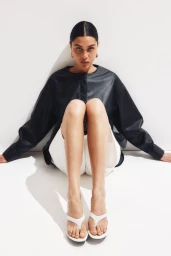 Model wearing a dark leather top and white shorts seated against a plain white studio backdrop for H&M.