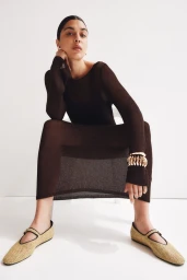 Model seated wearing a dark brown knit dress, chunky gold bracelets, and woven Mary Jane shoes against a white background